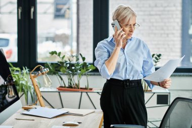 Kısa saçlı orta yaşlı iş kadını. Ofiste cep telefonu ve kağıtlarla birden fazla iş yapıyor..