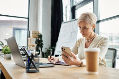 Orta yaşlı, kısa saçlı bir iş kadını, modern ofis ortamında dizüstü bilgisayar ve cep telefonu kullanarak bir masada oturuyor..