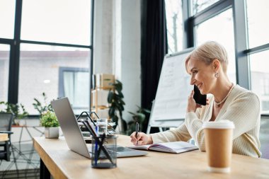 Kısa saçlı orta yaşlı iş kadını telefonla konuşuyor ve modern ofiste dizüstü bilgisayar kullanıyor..