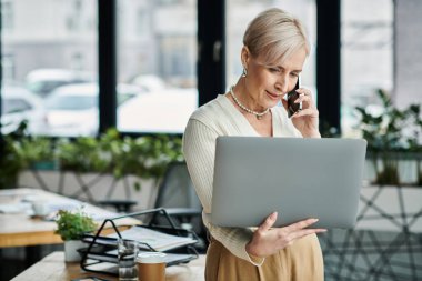 Modern ofis ortamında dizüstü bilgisayarla yazarken kısa saçlı orta yaşlı bir iş kadını cep telefonuyla konuşuyor..