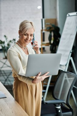 A middle-aged businesswoman with short hair simultaneously talks on a cell phone and works on a laptop in a modern office setting. clipart