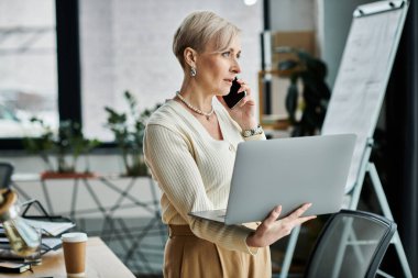 A middle-aged businesswoman with short hair talks on a cell phone while holding a laptop in a modern office. clipart