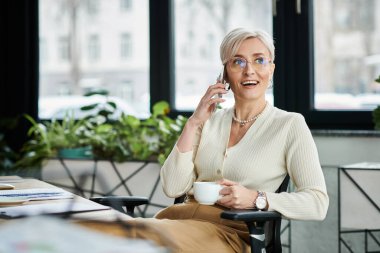 Orta yaşlı, kısa saçlı bir iş kadını, modern bir ofisteki cep telefonuyla konuşurken bir sandalyeye oturup odaklanır..
