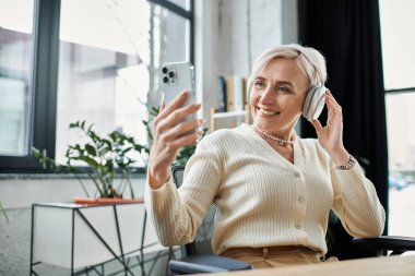 Orta yaşlı, kısa saçlı bir iş kadını bir masada oturmuş, modern ofis ortamında cep telefonuyla konuşuyor..