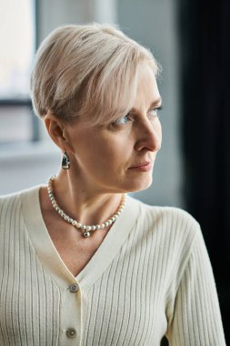 A middle-aged businesswoman with short hair wears a white sweater and pearls in a modern office setting. clipart