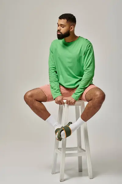 stock image Handsome African American man in fashionable attire striking a pose on a vibrant backdrop while sitting on top of a white stool.