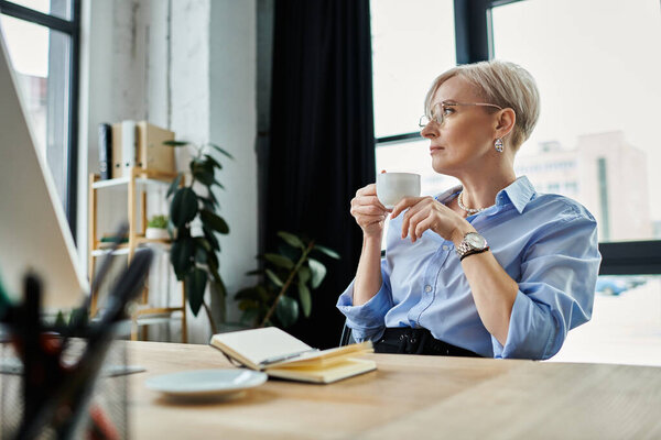 Деловая женщина средних лет с короткими волосами сидит за столом, держа чашку кофе.