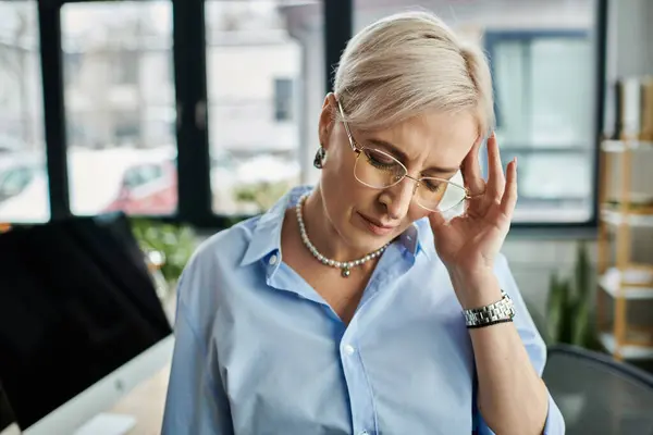 stock image Middle aged businesswoman with short hair experiencing menopausal symptoms, holding head in distress in office.