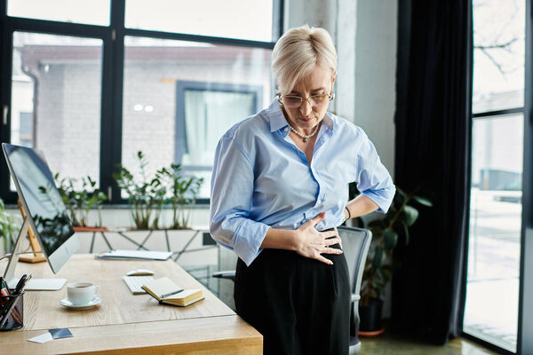 Деловая женщина средних лет, держа желудок в дискомфорте, стоя перед столом в офисе.