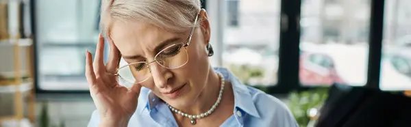 stock image Middle-aged businesswoman in blue shirt holding head with discomfort, facing challenges during menopause in the office.