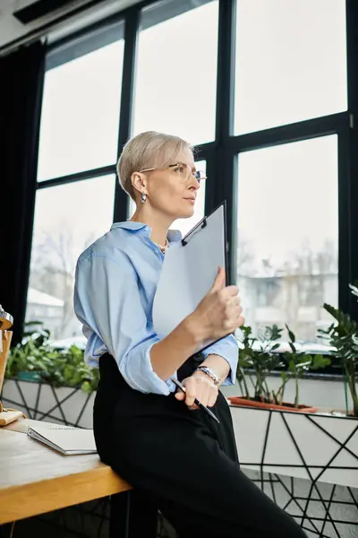 stock image A middle-aged businesswoman with short hair in a blue shirt and black pants diligently working at her desk