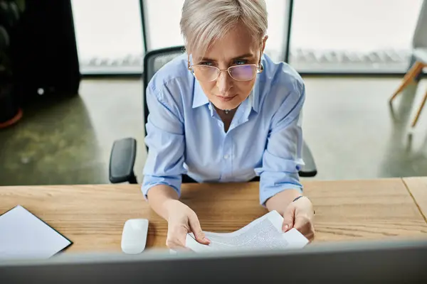 stock image Middle-aged businesswoman with short hair focusing on a piece of paper at her office desk during menopause.