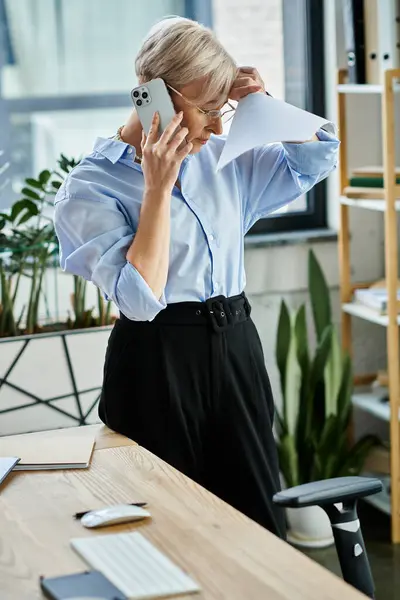 stock image A middle aged businesswoman with short hair, during menopause, stands in an office engaged in negotiations on a cell phone.