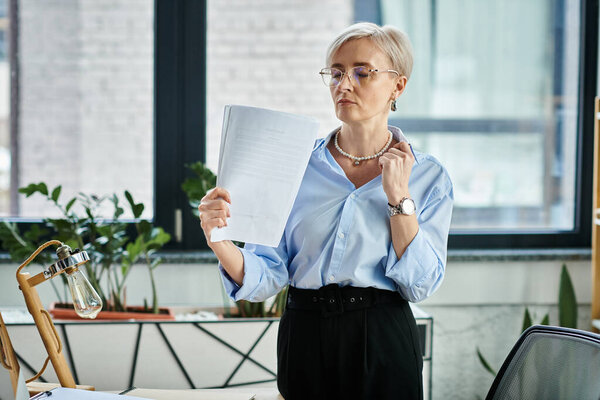 A middle-aged businesswoman with short hair, deep in thought, holding a piece of paper in her hands in an office setting.