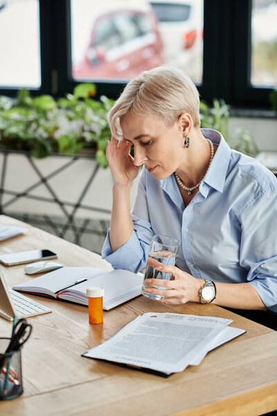 Деловая женщина средних лет с короткими волосами сидит за столом, глубоко задумавшись, пока держит стакан воды.