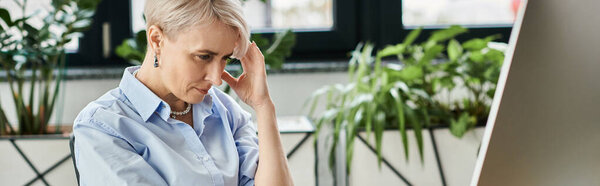 Middle-aged businesswoman sitting at computer desk, holding head in discomfort during menopause symptoms.