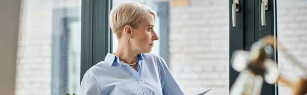 stock image Middle-aged businesswoman holding clipboard, standing by window in office.