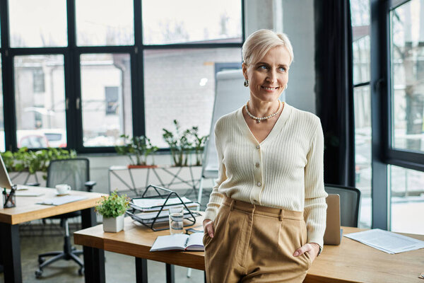 Деловая женщина средних лет с короткими волосами уверенно стоит перед столом в современном офисе.