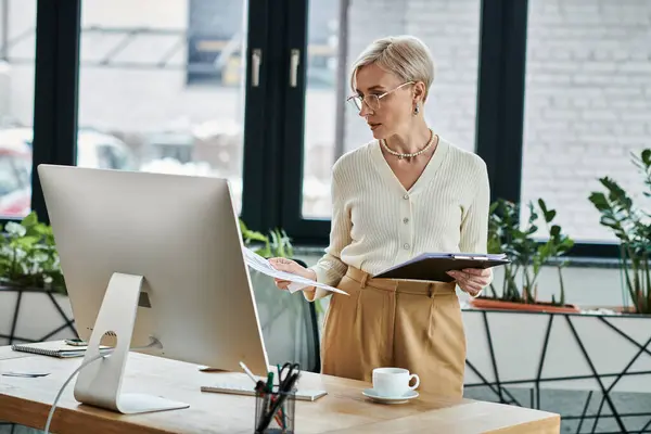 stock image A middle-aged businesswoman with short hair stands attentively in front of a computer in a modern office setting.