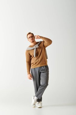 A handsome man in a brown sweater and grey pants striking a stylish pose. clipart