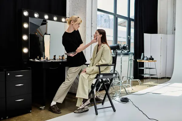 stock image A woman sits in a chair, as a skilled makeup artist works on her face in front of a mirror.