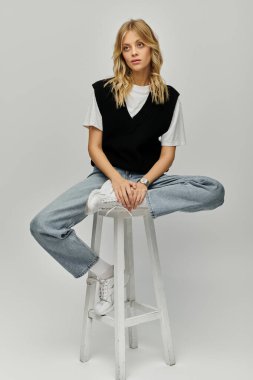 A stylish young woman with blonde hair sits elegantly atop a modern stool in casual attire against a solid grey backdrop. clipart