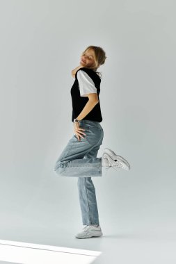 Sarı saçlı genç bir kadın, siyah gömlekli ve kot pantolonlu gri arka planda poz veriyor..