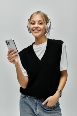 Sarı saçlı, kulaklık takan şık bir genç kadın gri bir arka planda cep telefonu tutuyor..