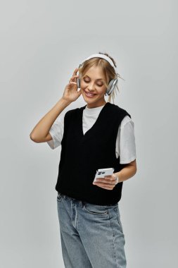 Şık giyinmiş, kulaklık takan, gri arka planda cep telefonuyla konuşan genç sarışın bir kadın..