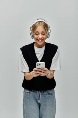Sarışın ve şık giyinmiş genç bir kadın cep telefonuna odaklanırken kulaklıkla müzik dinliyor..