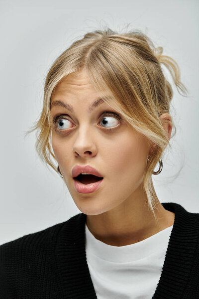 A young woman with blonde hair and stylish attire, looking surprised against a grey backdrop.