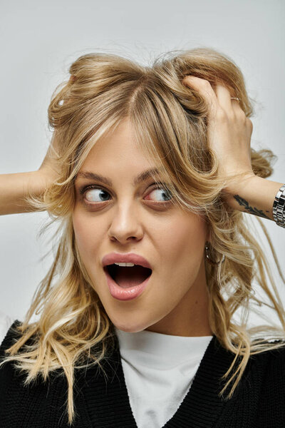 Молодая женщина в стильной одежде, держащая светлые волосы перед лицом на сером фоне.