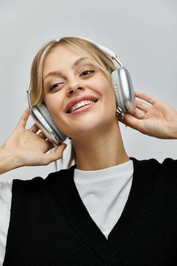 Young blonde woman in stylish attire listening to music with headphones on against grey background. clipart