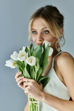 A beautiful young blonde woman gracefully holds a bouquet of white flowers against a soft grey background. clipart