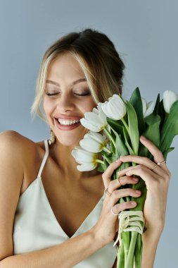 A young woman with blonde hair gracefully holds a bouquet of white flowers against a grey backdrop. clipart