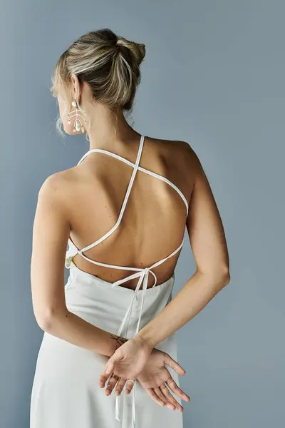 stock image A blonde woman exudes elegance in a white dress with a cross back design on a grey background.