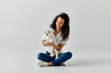 Bir kadın huzur içinde yerde oturuyor, beyaz köpeğini nazikçe kollarına alıyor..