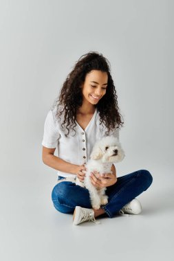 Genç bir kadın yerde oturuyor, nazikçe sevgili beyaz Bichon Frise köpeğini tutuyor.
