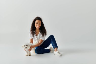 Genç bir kadın yerde oturuyor, yavaşça küçük Bichon Frise köpeğini tutuyor..