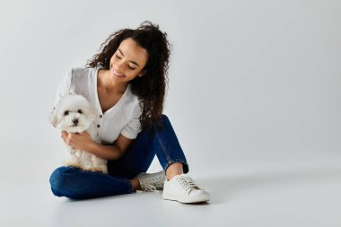 Yerde oturan bir kadın, nazikçe kollarında beyaz bir köpek tutuyor..
