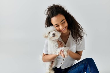 Genç bir kadın yerde oturuyor, kollarında beyaz bir bizon friz köpeği tutuyor..