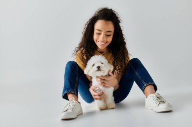 Genç bir kadın sakince yerde oturuyor, pofuduk beyaz bir Bichon Frise 'ye sarılıyor..