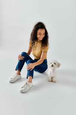 Bir kadın yerde pofuduk beyaz bir köpeğin yanında oturuyor..