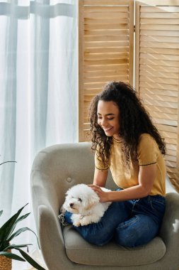 Young woman sitting on chair holding her bichon frise dog. clipart