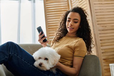 Kadın koltukta gevşiyor, tüylü beyaz bir köpek tutuyor ve telefonu kontrol ediyor..