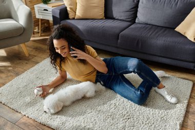 Yerde oturan genç bir kadın cep telefonuyla bichon frizini yakınlarda kullanıyor..