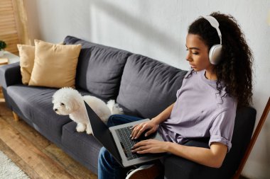 Kanepede dizüstü bilgisayarı olan bir kadın, yanında da bichon frise köpeği var..