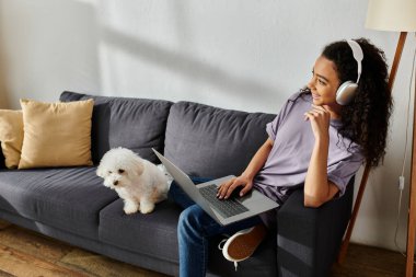 Kanepede dizüstü bilgisayarıyla oturan bir kadın, köpeğini okşuyor..