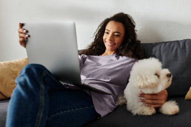 Genç bir kadın dizüstü bilgisayarıyla kanepede dinleniyor. Yanında da sevimli köpeğiyle birlikte..