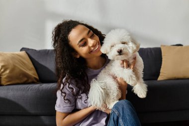 Bir kadın kanepeye uzanır, beyaz Bichon Frise köpeğini tutar..